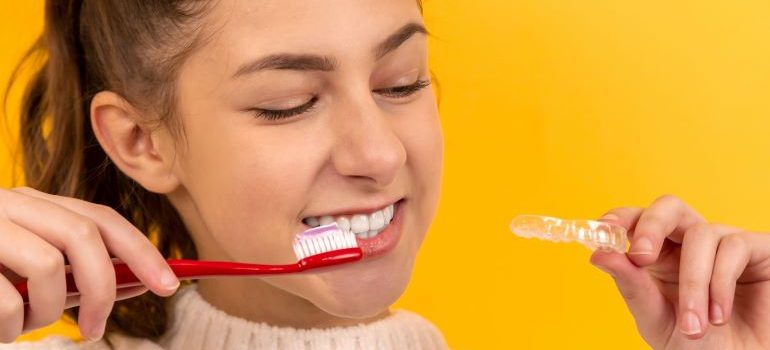 Girl brushing her teeth while holding a transparent teeth aligner in her hand.